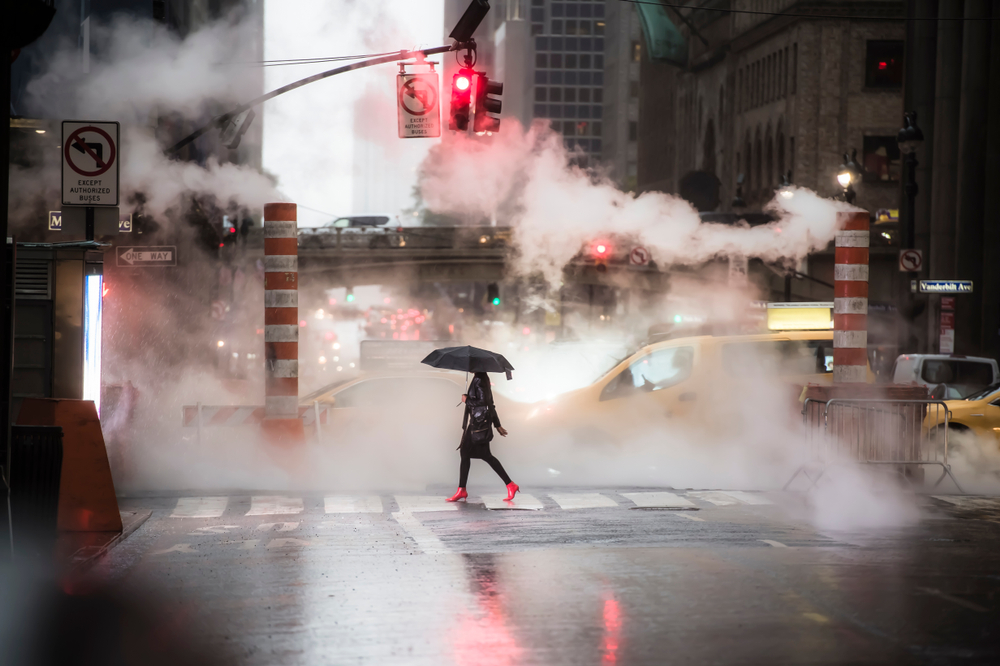 A person walks across a street holding an umbrella on a foggy, rainy day in the city. Steam rises from the ground, and traffic, including yellow cabs, is seen in the background under a red traffic light.