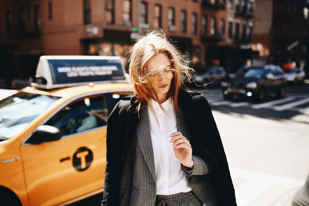 A woman dressed in a gray suit and black overcoat walks in the city on a windy day. Her hair is tousled by the wind as she strolls past a yellow taxi in the background.