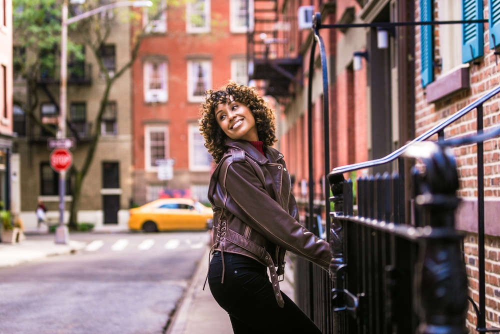 A woman in a leather jacket leans against a railing on a quiet city street, smiling brightly. A yellow taxi is parked in the background, and brick buildings with fire escapes frame the urban scene.