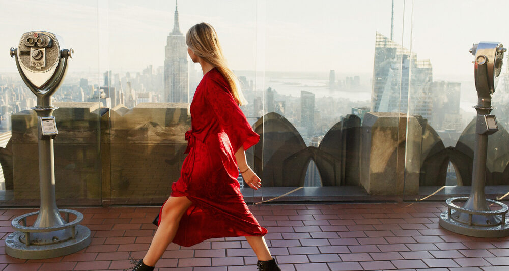 woman wearing a red dress walking with NYC skyline in the background