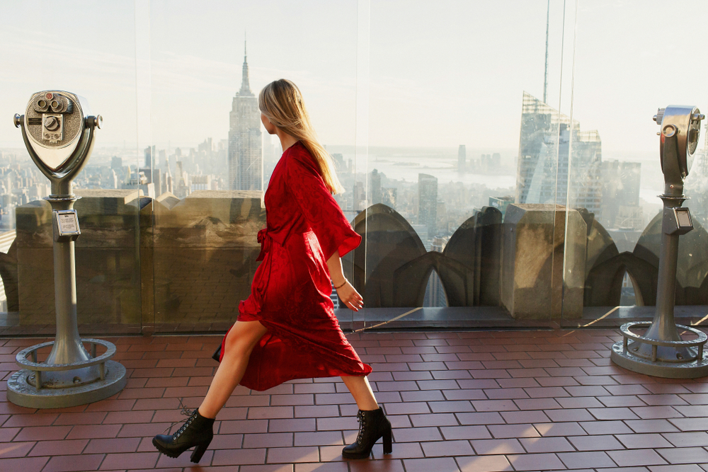 A woman in a flowing red dress walks across an observation deck with the New York City skyline, including the Empire State Building, in the background. Coin-operated binoculars are stationed on either side, adding a classic tourist vibe to the stylish moment.