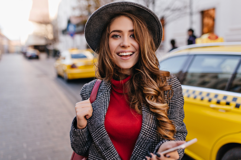 A smiling woman in a gray hat and checkered coat holds her phone and red bag while standing near yellow taxis. The setting is bright and cheerful, capturing her casual and happy mood.