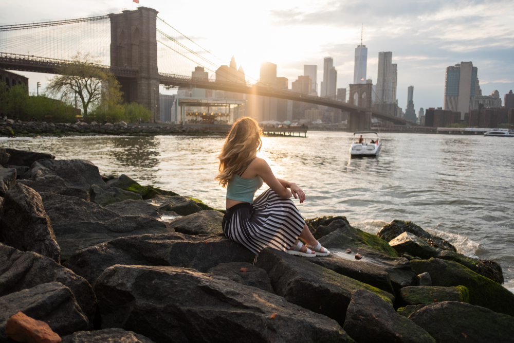 A woman sits on rocks near the waterfront with the Brooklyn Bridge and city skyline in the background at sunset. The scene is peaceful, with soft sunlight illuminating the water and the cityscape.