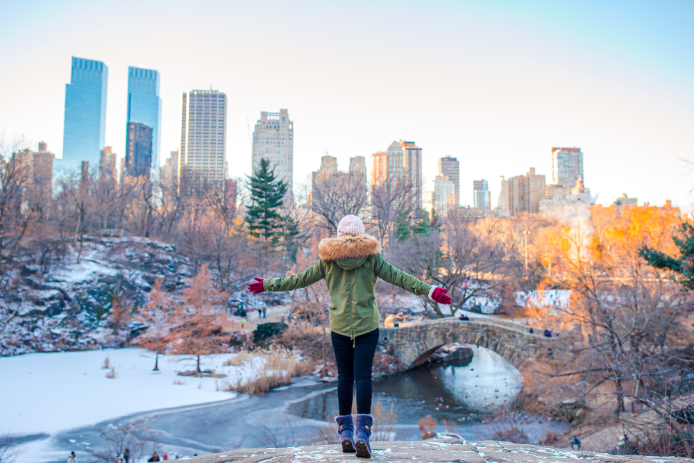 A woman stands with her arms open wide, facing the New York City skyline from Central Park during the winter. The scene features snowy trees, a frozen lake, and towering buildings in the background, evoking a sense of freedom and tranquility.