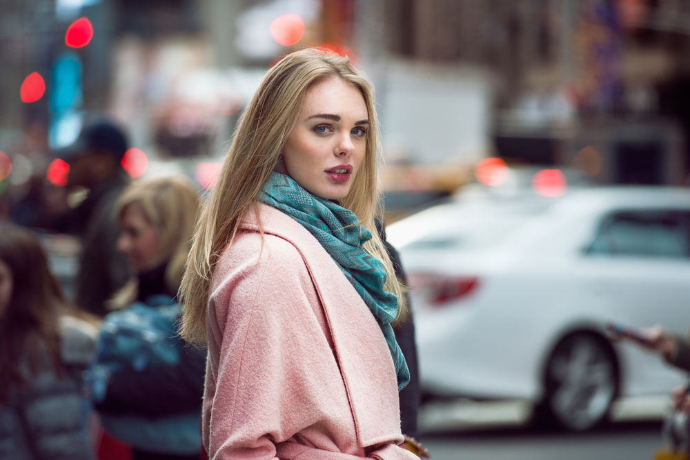 A woman wearing a pink coat and teal scarf looks over her shoulder on a busy city street. The blurred background includes pedestrians and cars, capturing a bustling, urban atmosphere.