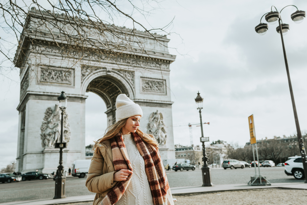 a fashionable woman is walking through the streets of Paris. She is wearing a stylish, chic outfit that blends with the Parisian street scene, exuding a sense of elegance. The background includes classic Parisian architecture, enhancing the city's timeless vibe