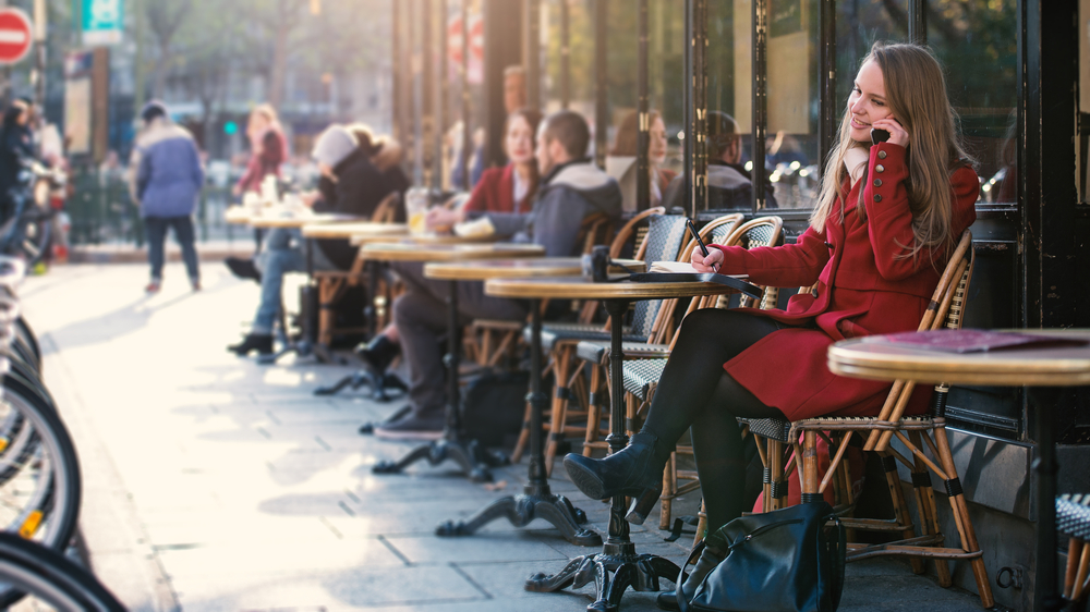 a woman sitting at an outdoor Parisian café in December. She is dressed warmly in a chic winter coat and scarf, enjoying the cozy ambiance typical of Paris in the colder months. The café's setting and street view capture the essence of a winter day in the city