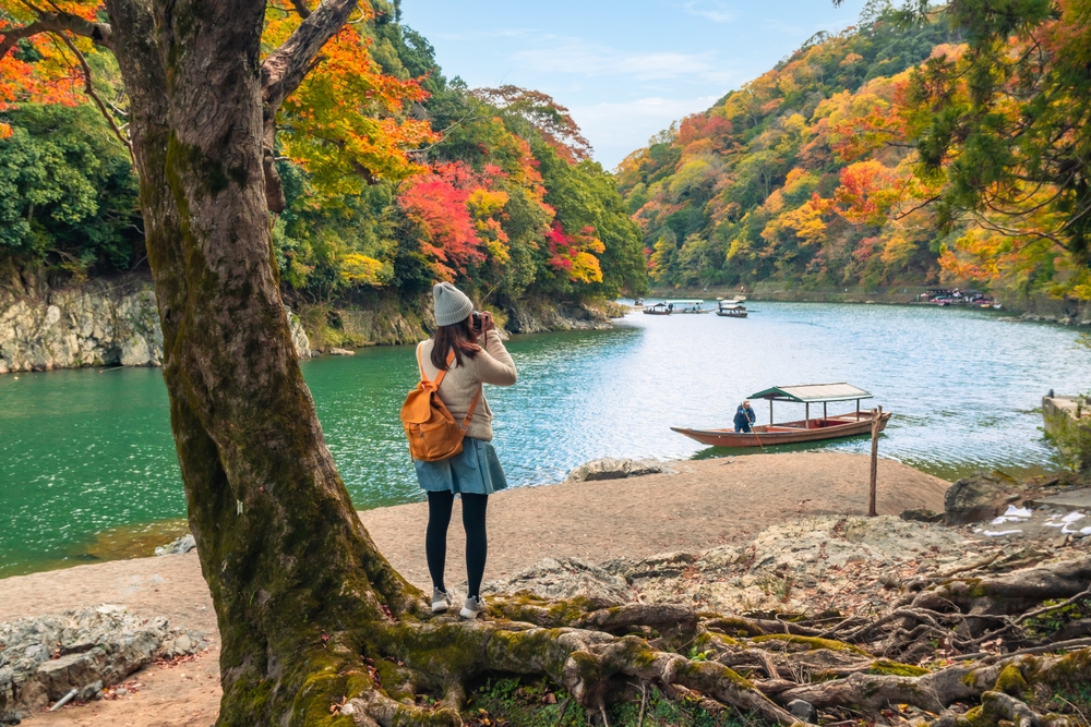 A solo traveler wearing a beanie and backpack photographs a serene riverside scene surrounded by vivid autumn foliage, with a traditional wooden boat on the water.