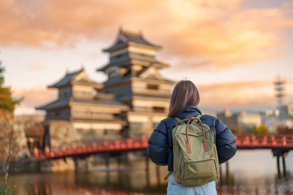 A traveler with a green backpack and puffer jacket stands on a bridge, gazing at a traditional Japanese castle at sunset, with the sky painted in warm hues.