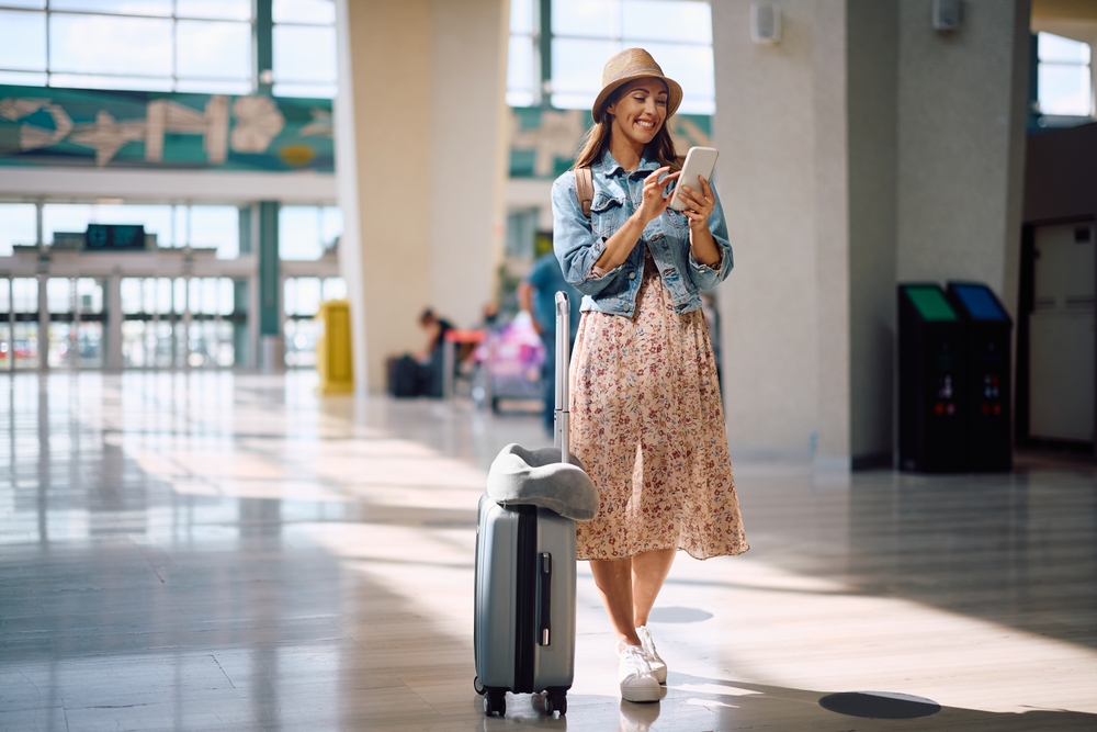 A cheerful woman in a floral dress, denim jacket, and sun hat stands in a bright airport terminal, looking at her phone beside a rolling suitcase.