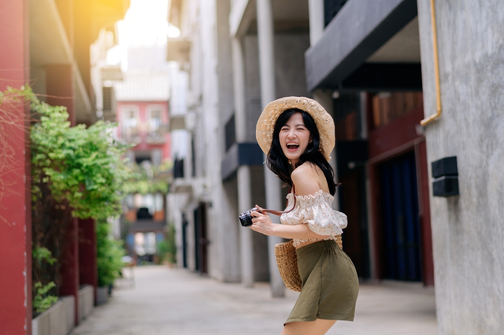 A smiling woman in a straw hat and crop top holds a camera in a modern, colorful alleyway, ready to capture the moment.