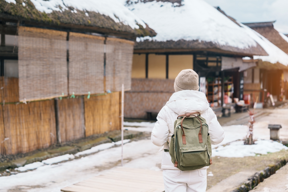 A traveler in a winter jacket and beanie with a green backpack explores a snowy traditional village with thatched-roof houses.