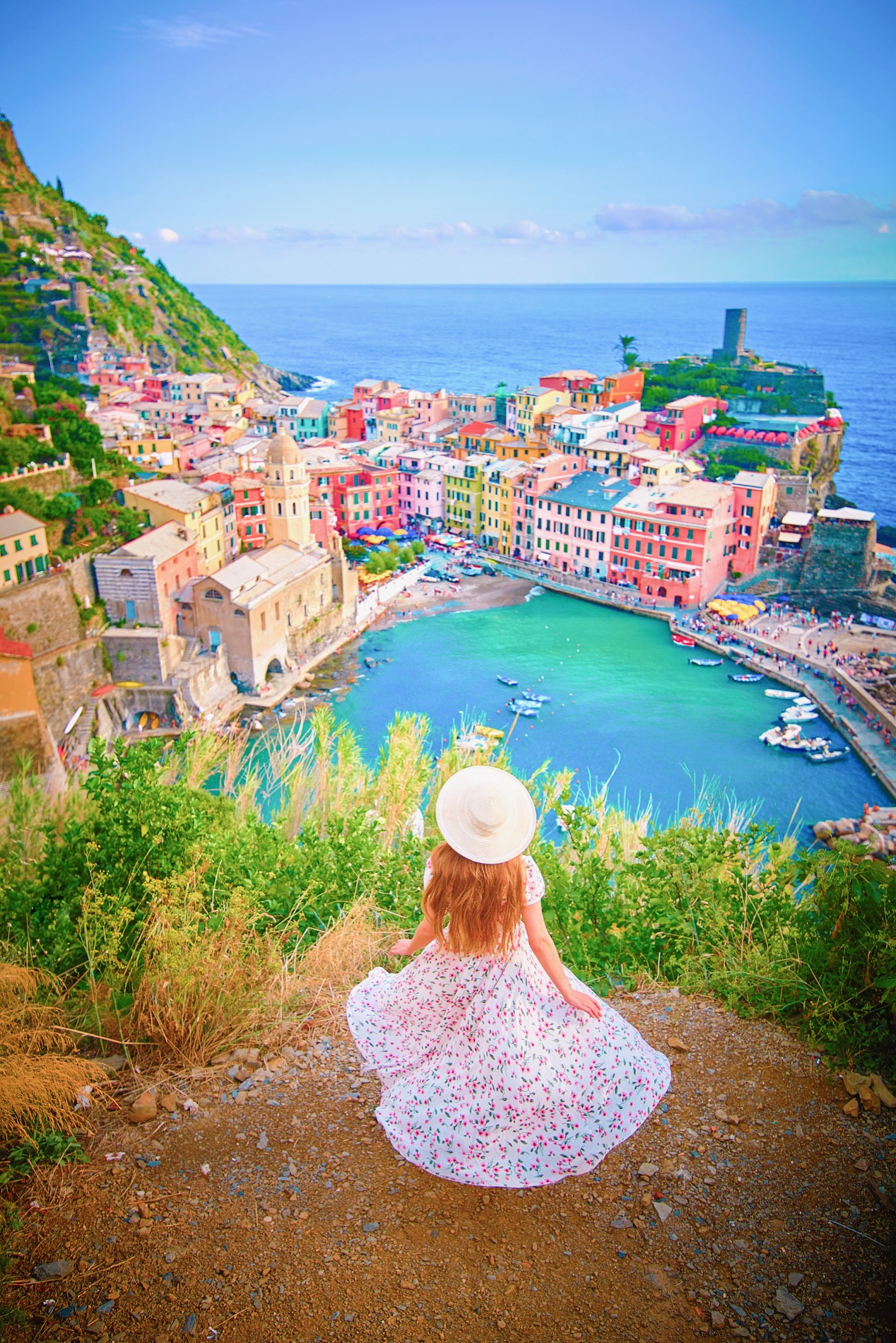 a girl in a sundress with hat standing with her back looking down on the colorful city of Cinque Terra