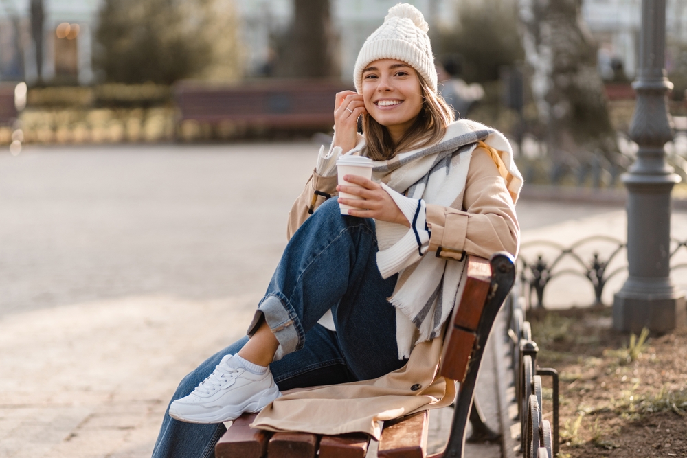 stylish woman walking in winter street wearing beige coat, knitted hat, scarf, smiling happy cold season fashion trend, drinking coffee. A perfect London January outfit