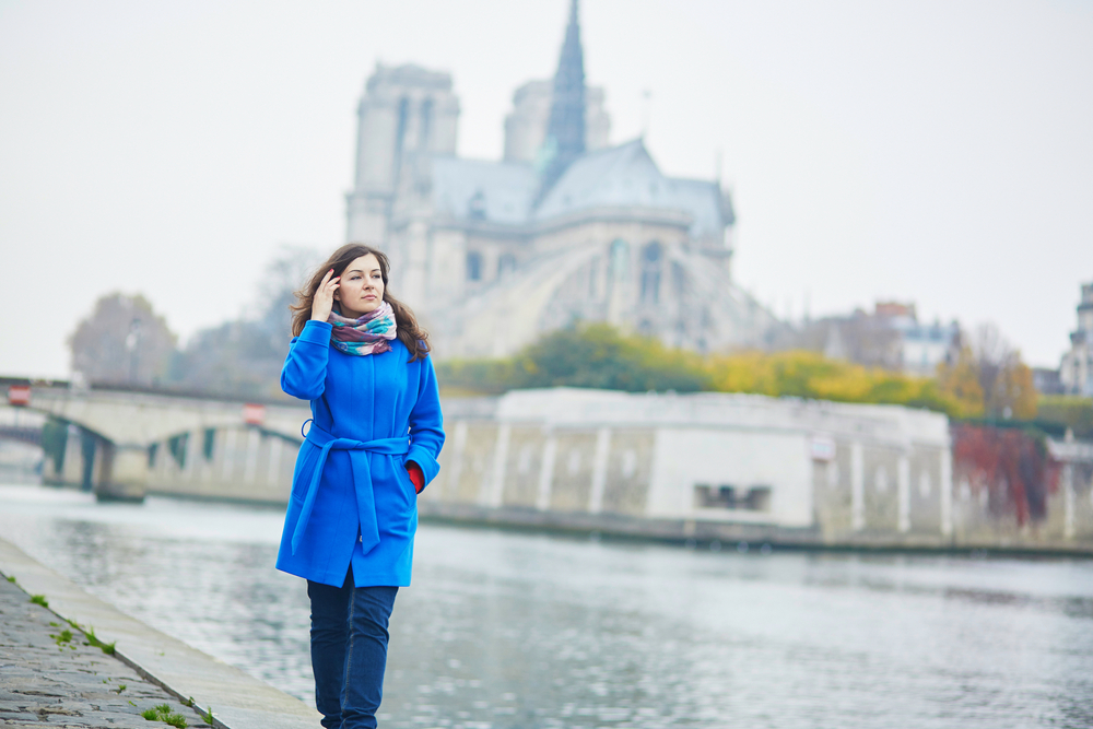 a woman is walking along the river in paris, she is wearing a wool coat and stylish scarf as she brushes her hair behind her ear