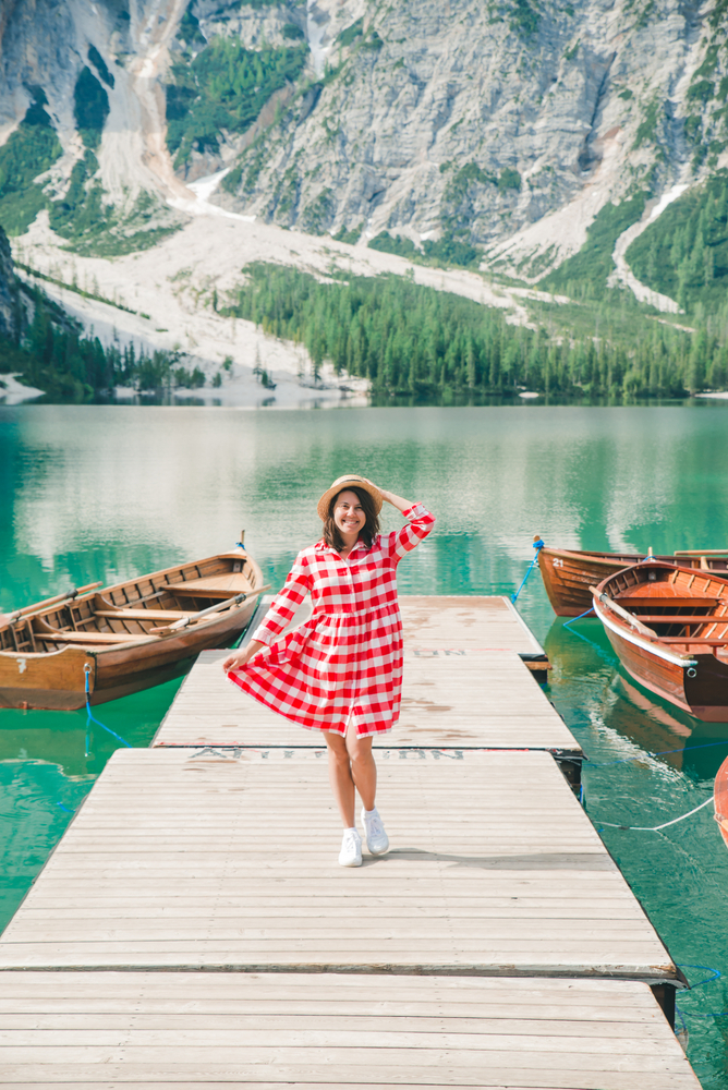 a woman in a red dress and white sneakers standing on a dock in the lake overlooking mountains