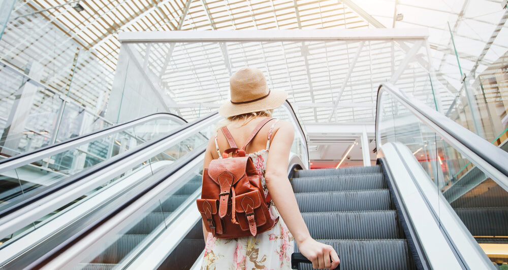 woman standing on escalator wearing hat and brown backpack at the airport in summer