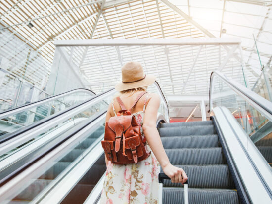 woman standing on escalator wearing hat and brown backpack at the airport in summer