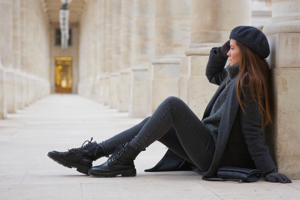 a woman is sitting down leaning up against a column while wearing combat boots, jeans, sweater, wool coat, and a beret 