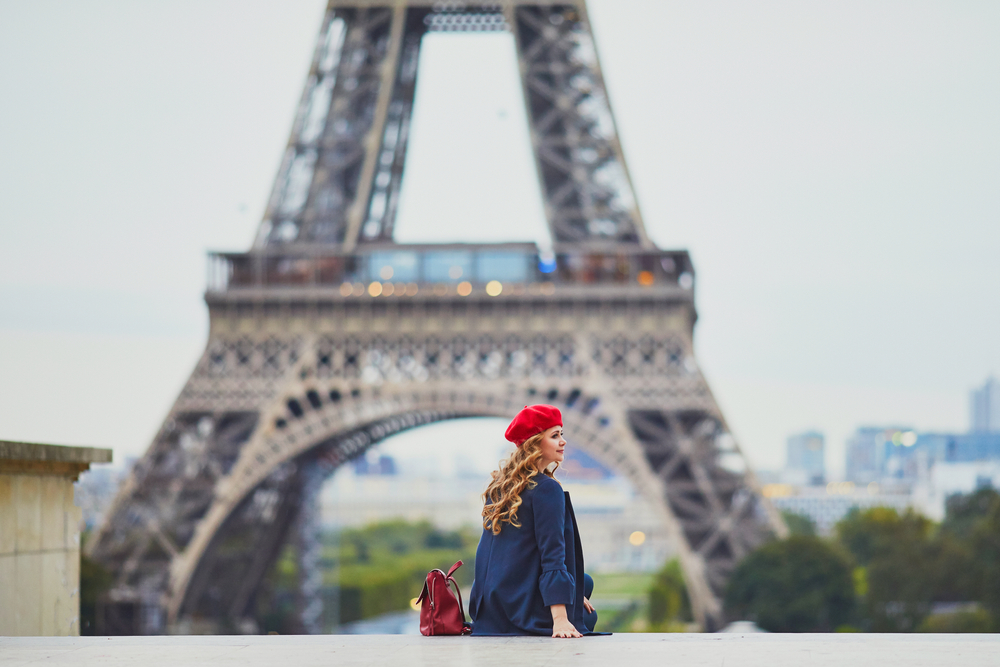 a woman sits on stairs in front of the eiffel tower. she is wearing a beret and a stylish wool coat, her bag sits next to her on the stair 