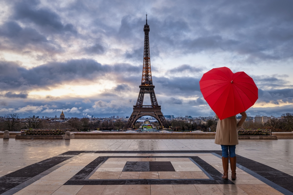 a woman stands in front of the eiffel tower holding a red heart shaped umbrella in February in paris 