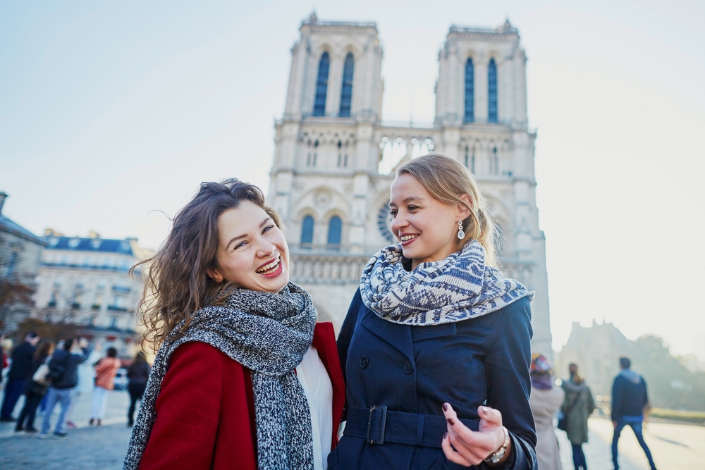 two women in winter outfits smiling in the streets of paris in february, they are both wearing scarves, people are walking in the background behind them 