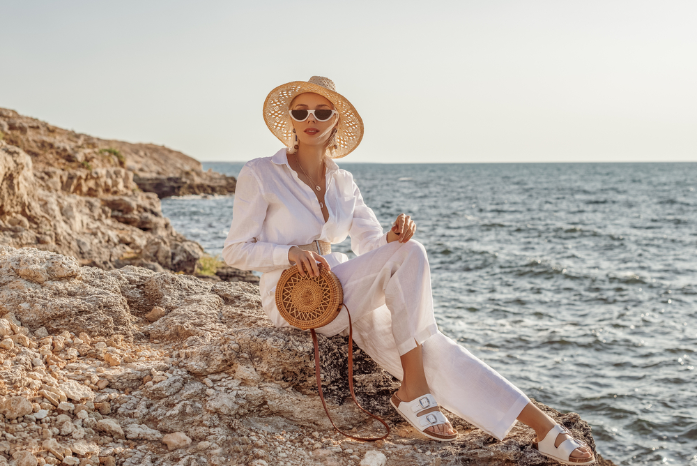 a woman sitting on rocks wearing white with straw hat and bags in front of the ocean 