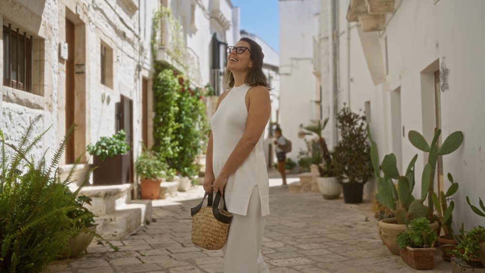 a girl in matching two piece outfit with straw bag  is ideal for what to wear to Italy in Summer standing in a cobblestone street surrounded by white buildings. 