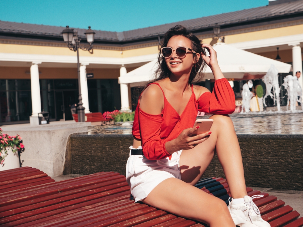 a girl in bold red top and white shorts and sneakers sitting poolside with phone in her hand 