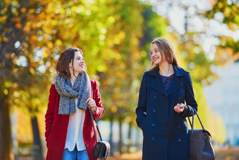 Two young woman walking in a fall park wearing light coats, scarves, and bags, staples of Paris Outfits in October.