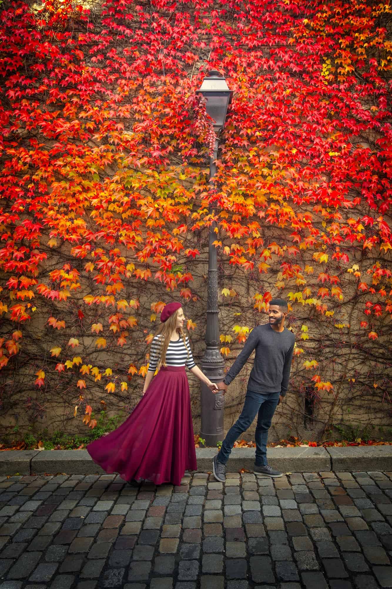 Woman in a long red skirt, black and white shirt, and red beret holds hands with a man wearing long=sleeved black shirt and black pants in front of a lamp post and orange and red ivy on a wall.