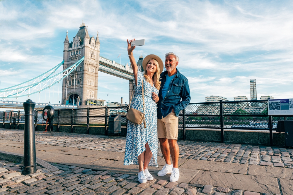 A couple taking a selfie in front of Tower Bridge along the river. The woman is wearing a white and blue dress with with sneakers, a great idea for what to wear in London in Summer.