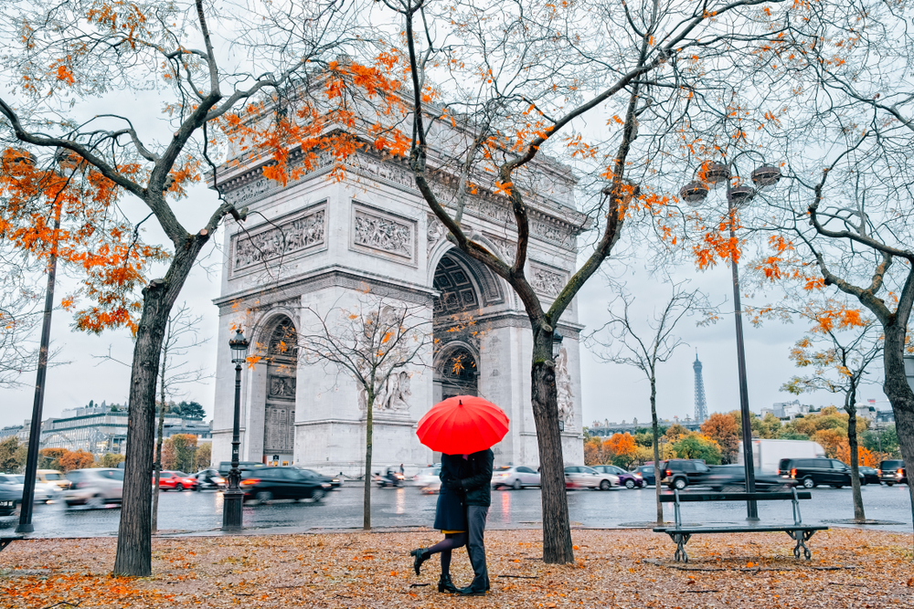 Couple under a red umbrella standing among fall foliage trees in front of the Arc de Triomphe.