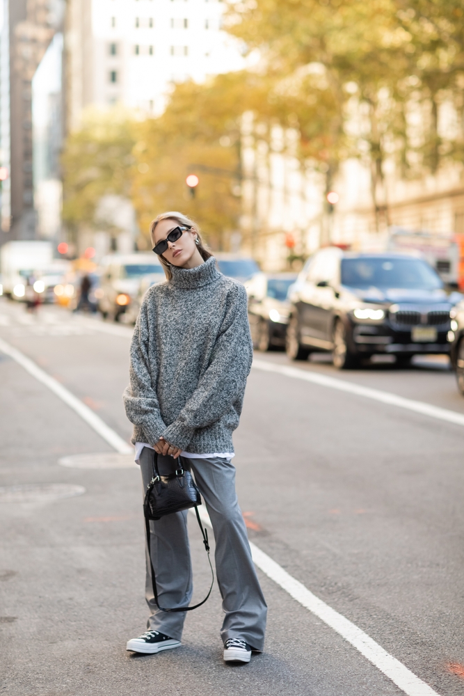 a woman is standing in the street wearing oversized pants and sweater with sneakers and a small bag in her hands, she also has on sunglasses, you can see cars on the road behind her