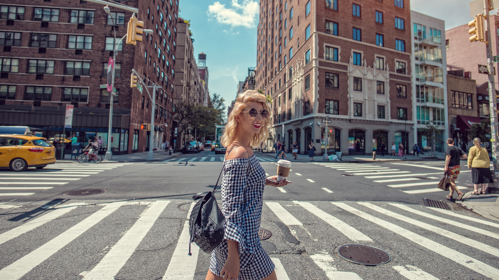 a woman is walking across a street  on a crosswalk, she is wearing a gingham dress with a black leather backpack, she has a phone and a to-go coffee cup in one hand, nyc outfits in spring