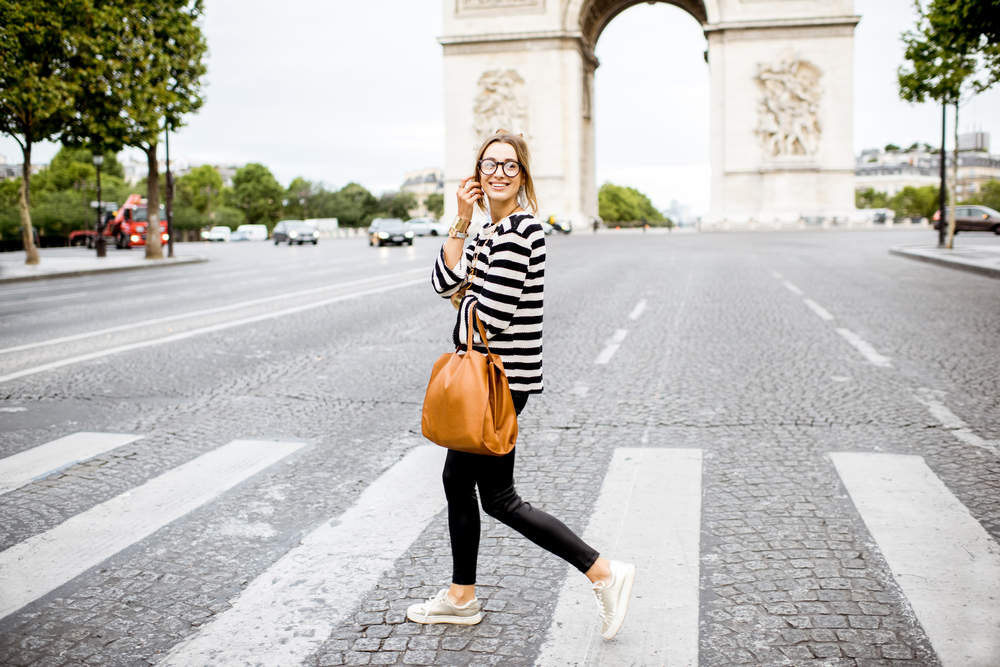 Woman with glasses crossing the street in front of the Arc de Triomphe wearing a long-sleeved black and white shirt, black pants, and white sneakers while carrying a brown bag.