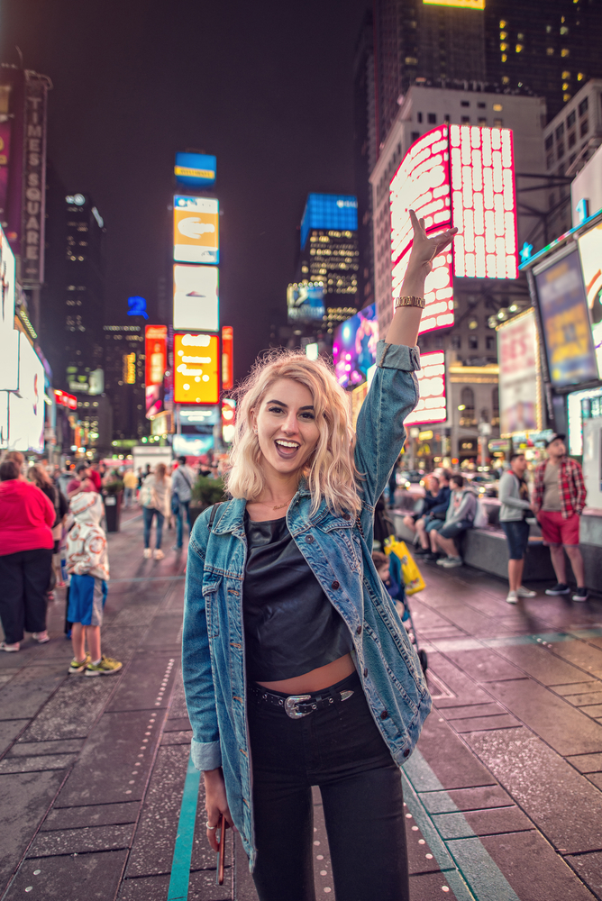 a woman is standing in time square wearing double denim with one hand raised in the air making a peace sign, billboards and people and buildings can be seen in the background 