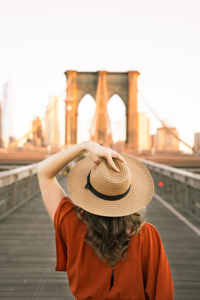 a woman is standing in front of a bridge her hair is all behind her and it is curled, she is wearing a hat and holding it on her head. nyc outfits for. spring 