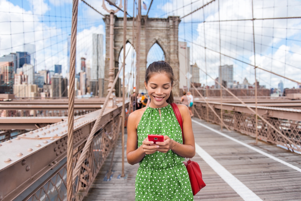 a woman stares at her phone on a bridge while wearing a flowy dress and a bright red bag on her shoulder, what to wear in nyc in summer 