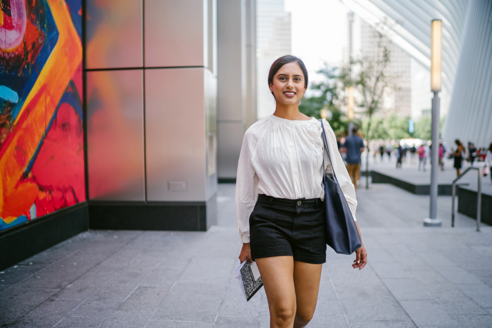 a woman in city street wearing a breezing white blouse with black shorts. she has a purse on one shoulder and is carrying a notebook.