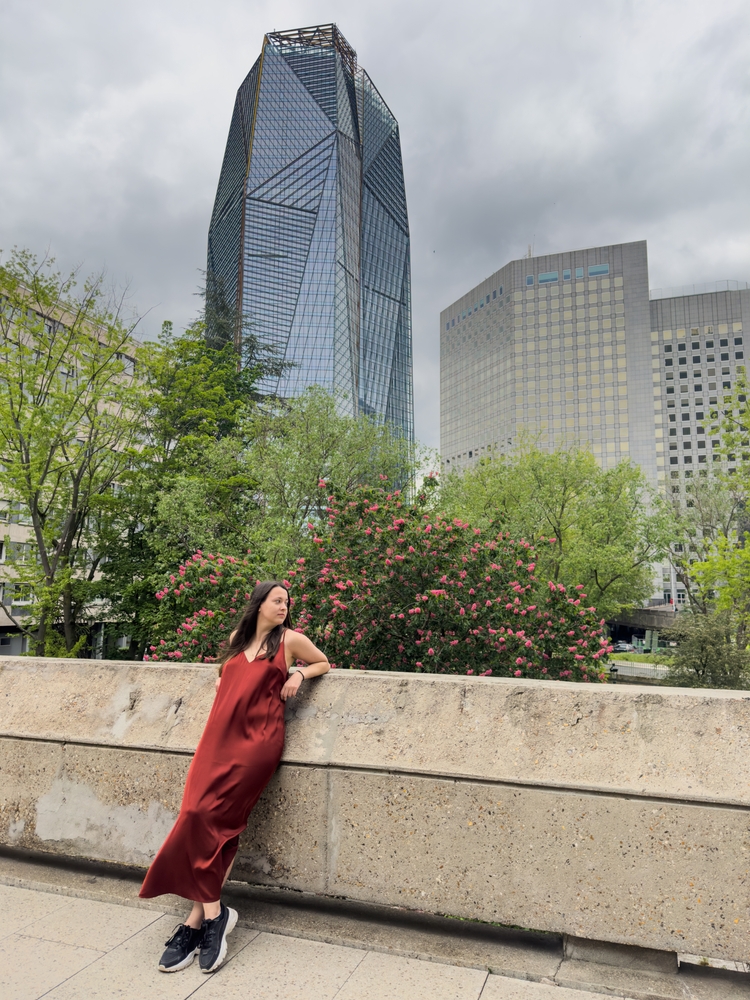 a woman leans up against a concrete wall wearing a long silk dress and sneakers, her hair is down, there are trees and buildings in the background 