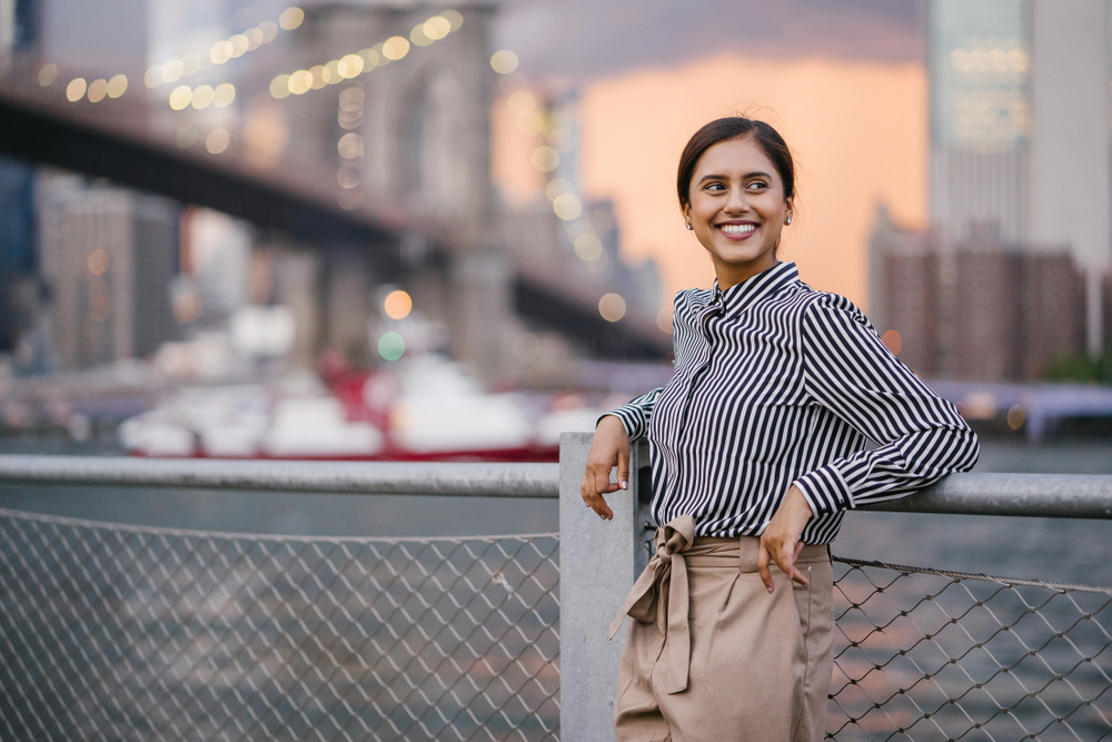 a woman stands in a striped button up short and paper bag trousers learning up against a fence next to a river, she is smiling into the distance 