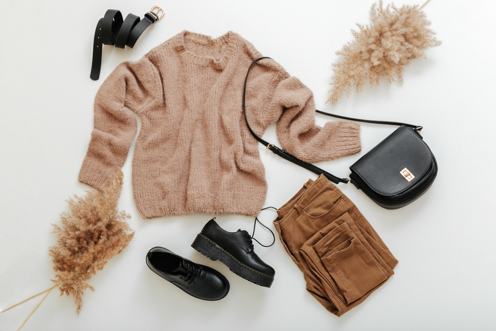 a sweater, shoes, pants, and a purse are laying on a white surface, there are two decorative plants around the outfit 