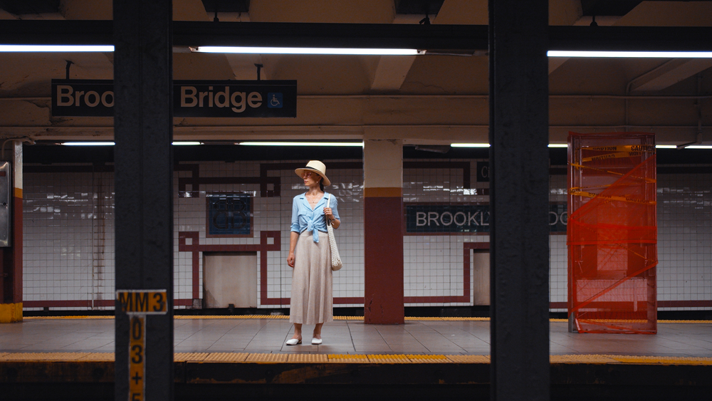 a woman dressed stylishly in a long skirt, blouse, and hat is standing in the subway waiting, nyc outfit 