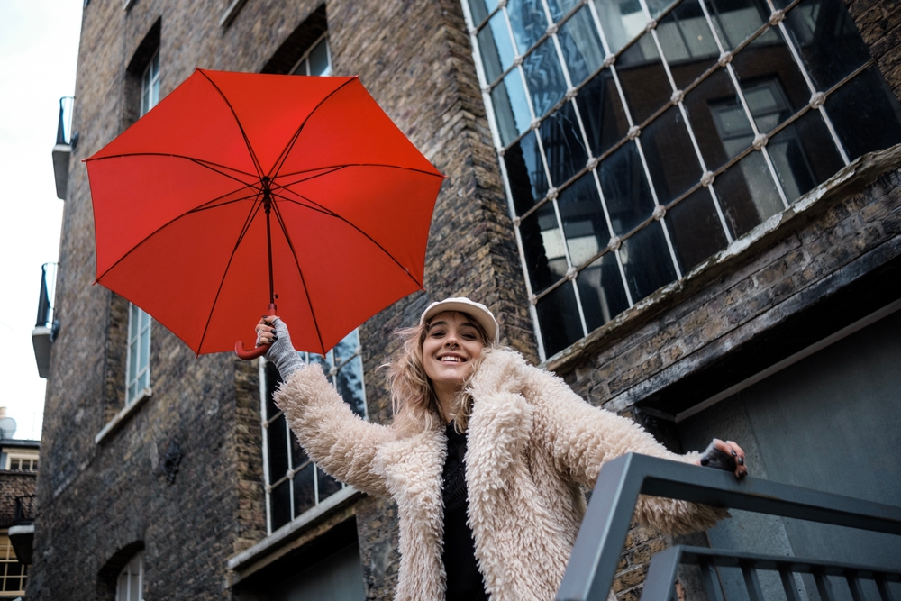 A women perches over the stairs of a building, wearing a fleece coat over her hand warmers, her long sleeve shirt and her hat. She holds a red umbrella and doesn't worry about what to wear in London in February. 