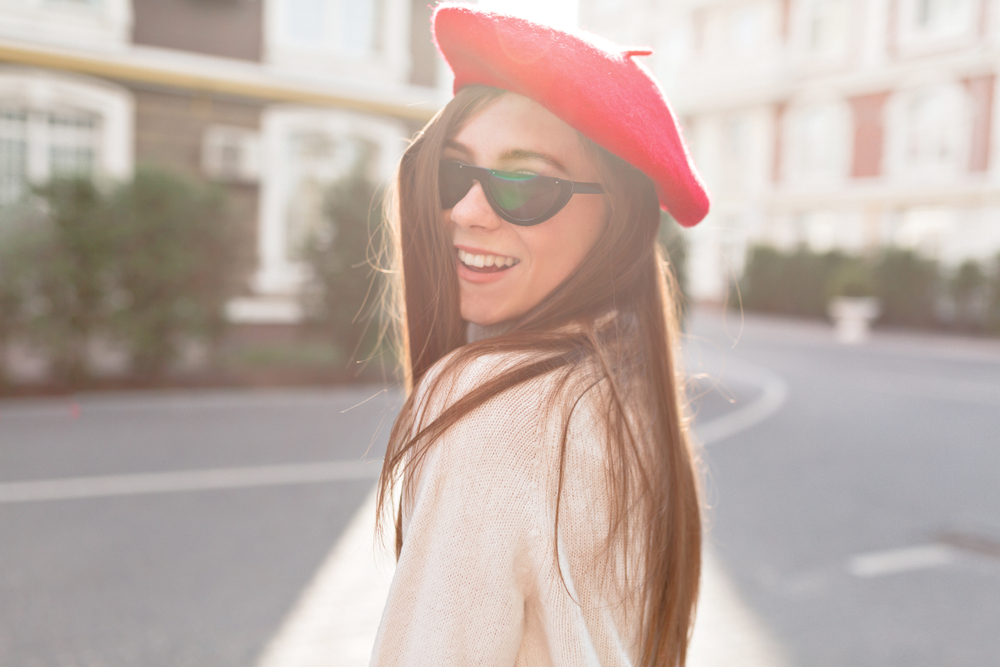 A young women looks over her shoulder and smiles in her white sweater, her red beret, and her fun sunglasses. 