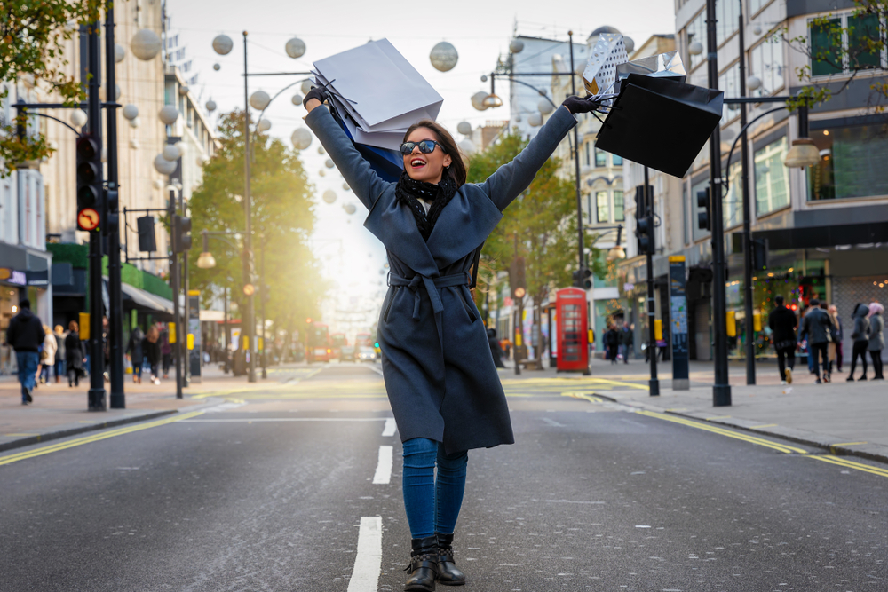 A women throws her shopping bags over her head as she walks down the street, enjoying her warmth in her jeans, boots, and navy trench coat. 