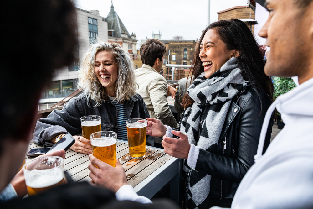 A group of friends wear plaid shirts, leather jackets, scarves and more as they gather around and drink at a pub, not worrying about what to wear in London in February. 