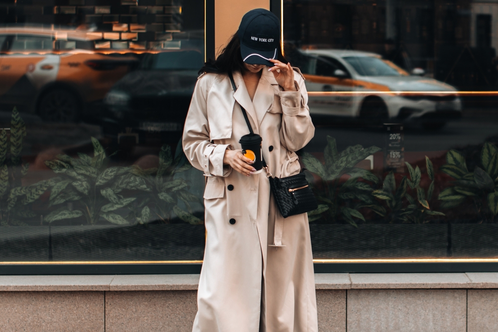 A women tips her hat while wearing a trench coat and drinking a coffee: she is staying warm in the city as cars pass by, not worrying about what to wear in London in February. 