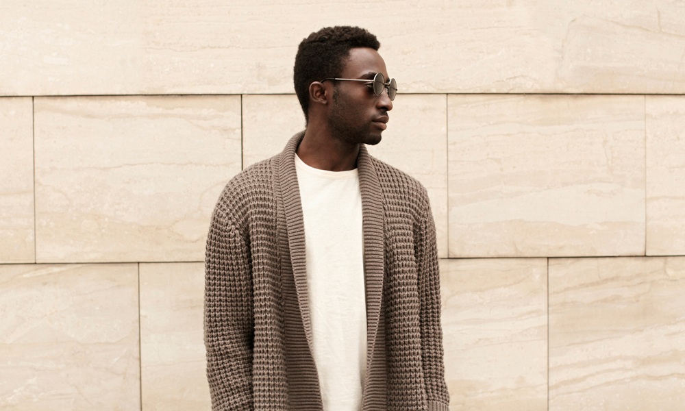 A young man stands in front of a museum wall in a heavy, knit cardigan and a long sleeve shirt: the perfect museum outfit. 